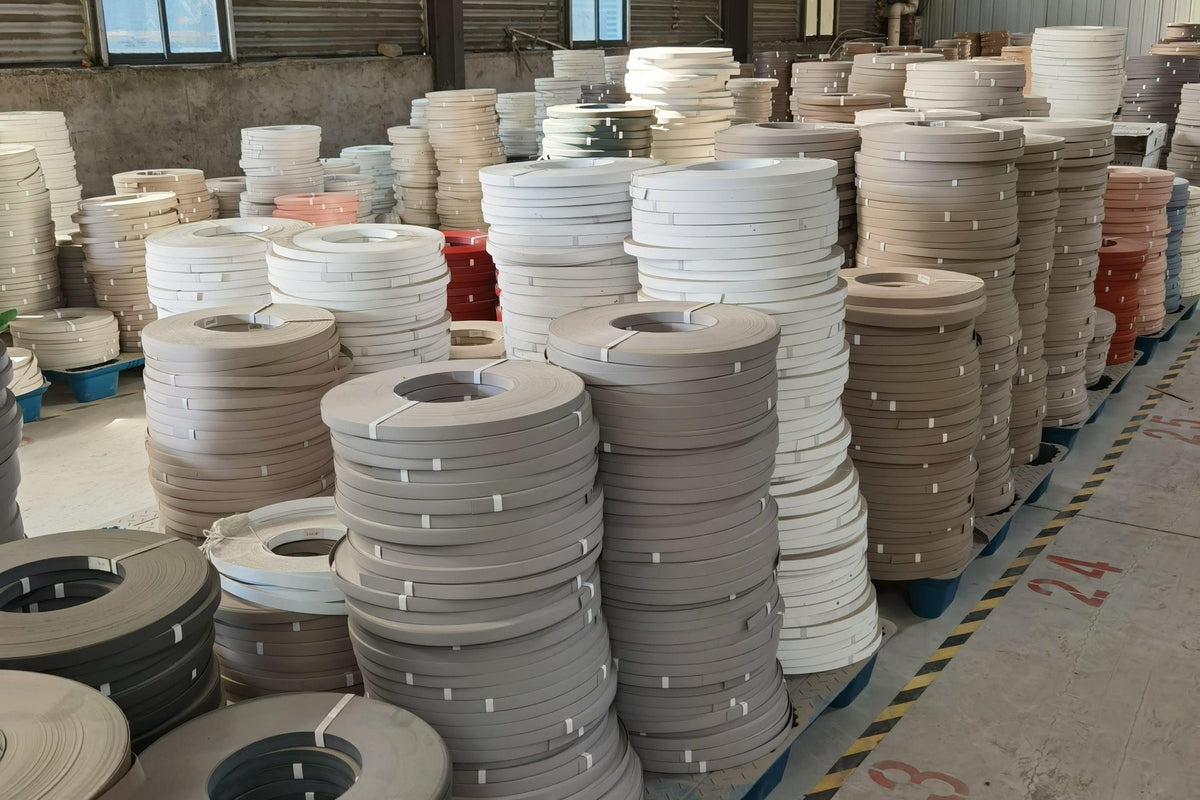 Stacks of edge banding rolls in white, grey, beige and darker tones on pallets in a warehouse